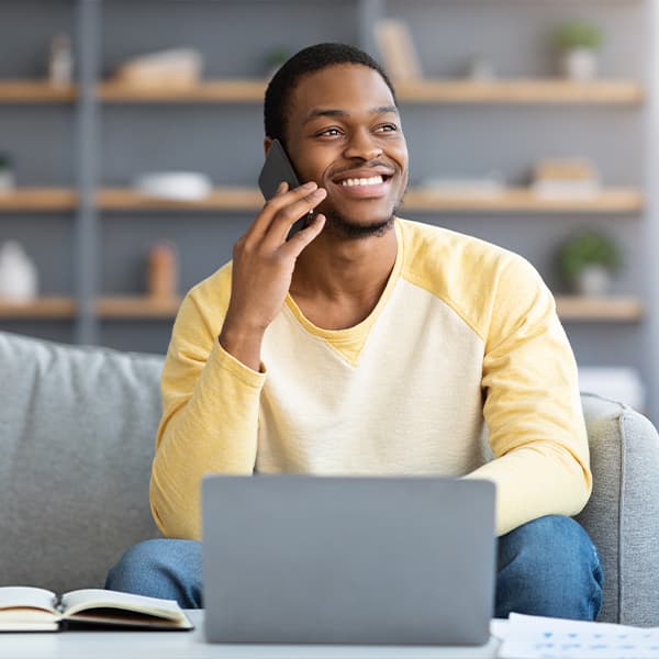 Joyful-black-guy-freelancer-having-phone-conversation-while-working-from-home